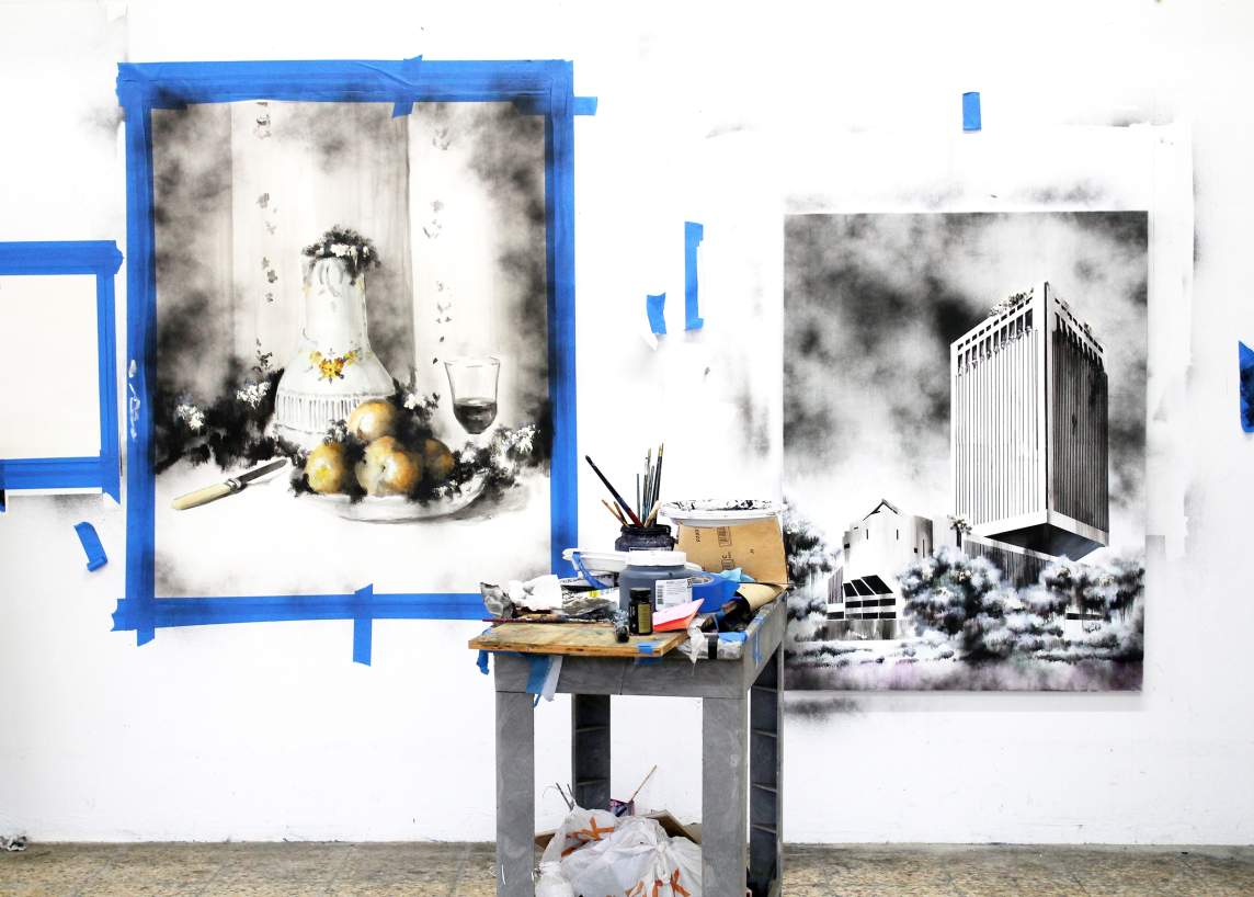 Art studio with two black-and-white paintings on the wall, one of a still life and the other of a modern building. A table with art supplies is in the foreground.