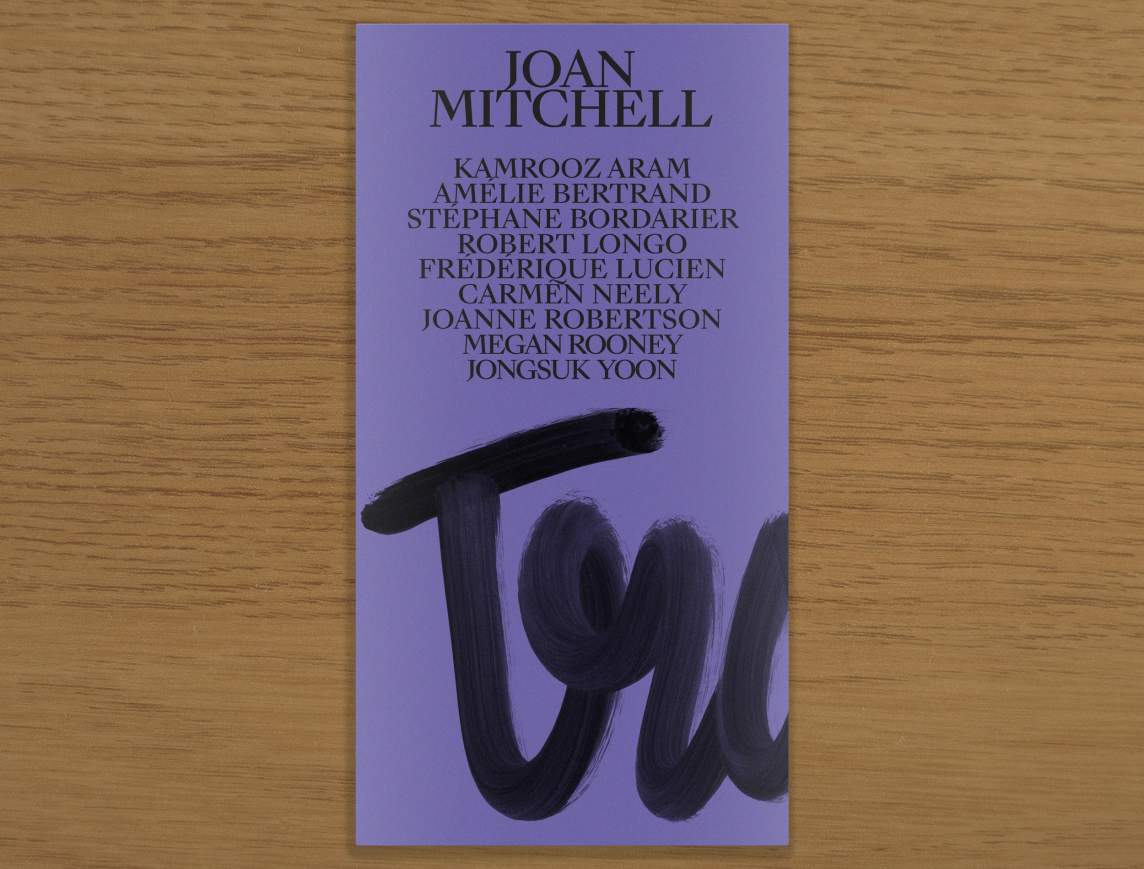 A narrow book with a purple cover lays on a wood table, the cover reads: “Joan Mitchell, Kamrooz Aram, Amelie Bertrand, Stephane Bordarier, Robert Longo, Frederique Lucien, Carmen Neely, Joanne Robertson, Megan Rooney, Jongsuk Yoon” Below the names is a partial word scrawled in vertical cursive reading “Tra”