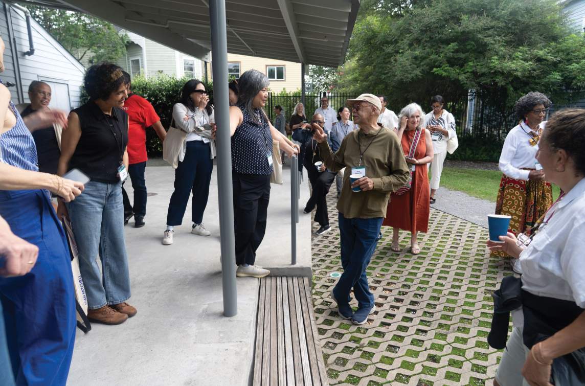 A group of over 20 people of various dress, age, and appearance mill about in conversation on a moss and stone patio and concrete porch with cantilever roof.