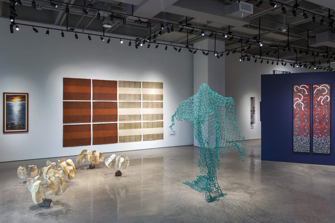 In a gallery space with white walls and polished concrete floors, several artworks are installed on the walls, and two sculptures stand in the foreground. The center sculpture is aquamarine blue netting suspended from the ceiling, evoking an invisible figure with arms outstretched beneath. To the left, are 6 low plant-like sculptures, with light papery leaves with writing on them extending from rocks. On the wall, is a vertical photograph of a sunset over water, and a grid of 6 orange and 6 ivory horizontal sheets of paper with fine lines drawn in horizontal stripes at the top and bottom of each.