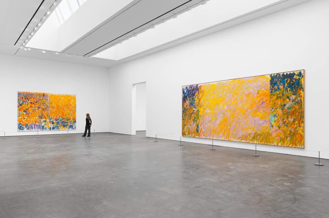 A person views two large abstract paintings by Joan Mitchell with dominant yellow and orange tones in a spacious, modern art gallery with white walls and a concrete floor. The painting on the left is a large diptych, and on the right is a quadriptych.