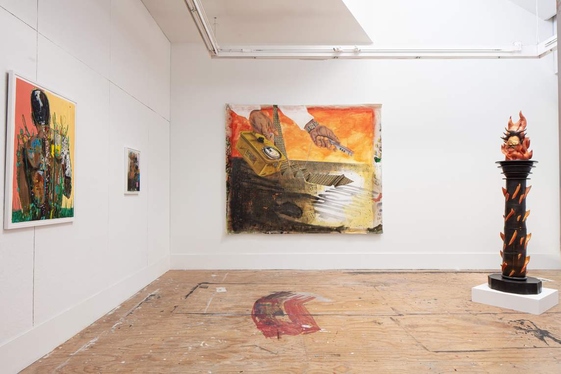 On the right, a columnar sculpture sits on the paint-splattered floor, of a man’s head with sunglasses at the top of a column, with dimensional flames coming out of the column and his head. On the white walls, three paintings hang, two smaller abstracted figurative works, and a larger painting with a man's hands taking radiation measurements, with a long tie hanging down, and words reading “FOR SALE.”
