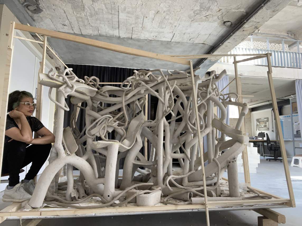 Linda Sormin sits to the side of a large sculpture sitting on a rolling cart. The sculpture is composed of bending rods and tendrils of white clay, intertwined throughout the form.