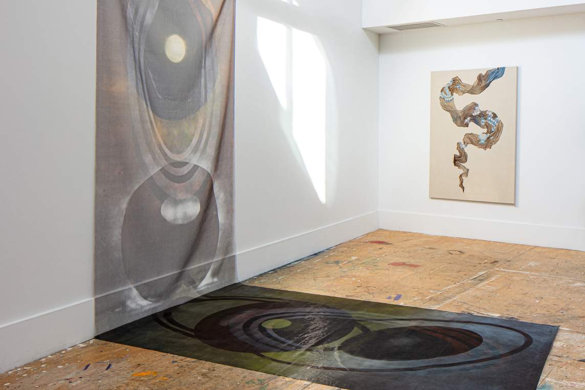 In an art studio with white walls and plywood floors, an abstract artwork with circle forms is pinned to the wall, with a similar fabric artwork resting on the floor in front of it. On the wall on the right, an abstract artwork hangs on the wall, with a skein of gauzy fabric swirling amidst a beige canvas.