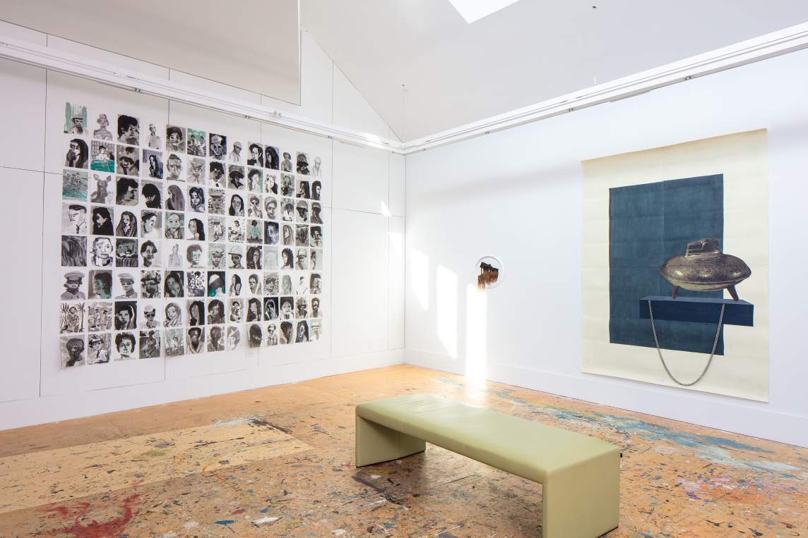 On the left wall, a grid of small pen and ink portraits of people are arranged with space in between them. On the right wall, a small wall-hung sculpture depicts an old house in a white frame with vegetative tendrils hanging down from it. At far right is a large mixed media work on paper with a central panel of dark blue, and a lidded metal pot resting on a blue shelf. A silver chain hangs on the surface of the painting from one edge of the painted shelf to the other.