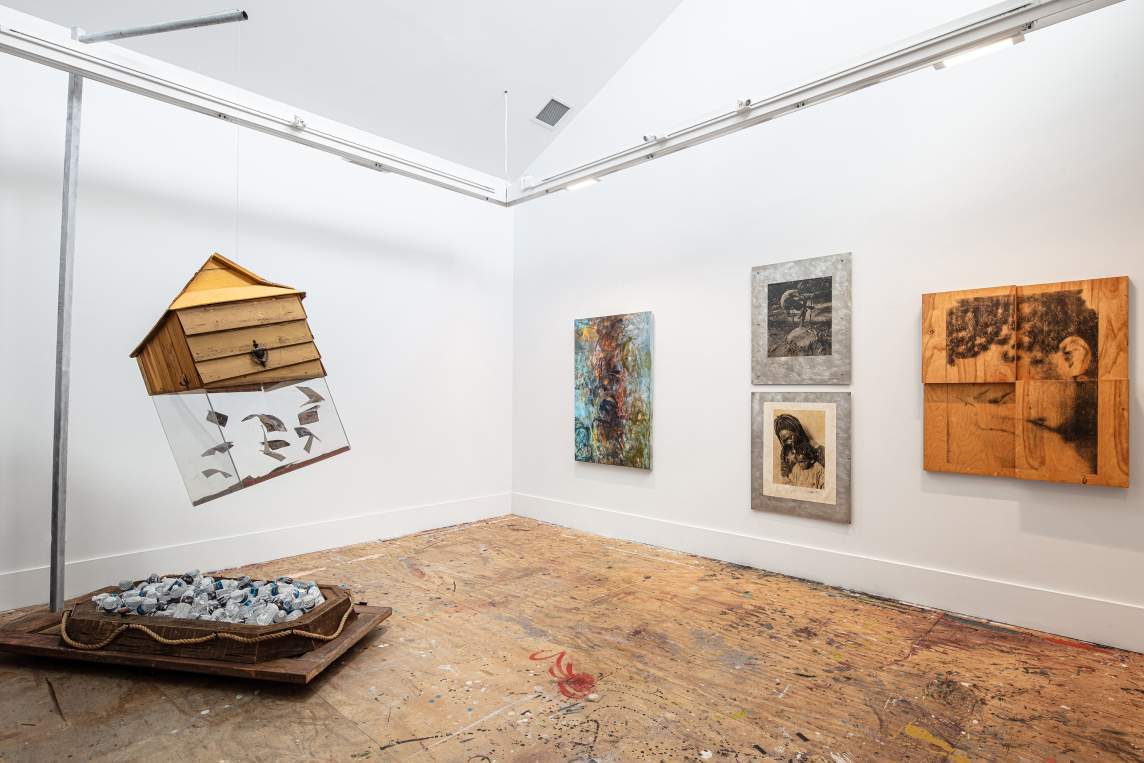 At left, a sculpture of a small house hangs from a metal stand. The plexiglass base of the house is filled with water-stained photographs. On the stand below, many water bottles rest in a small boat form. On the opposite wall are an abstract painting, with a dark central form and swirling brushstrokes, hung next to three portraits by Clifton Faust. One depicts an African-American mother and child, another depicts a portrait of an African-American woman etched on a wooden board.