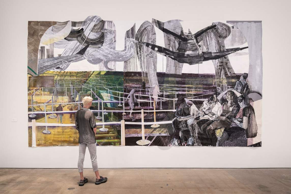 In an art gallery, a person with light skin and short blonde hair looks at a larger-than-life artwork, hung on a white wall. The piece, “In Between the Lines”, is a large painting with a group of figures sitting and huddled together at the bottom right corner, and beyond them a labyrinth of line stations. Above the scene are undulating swirls and airplane wings.