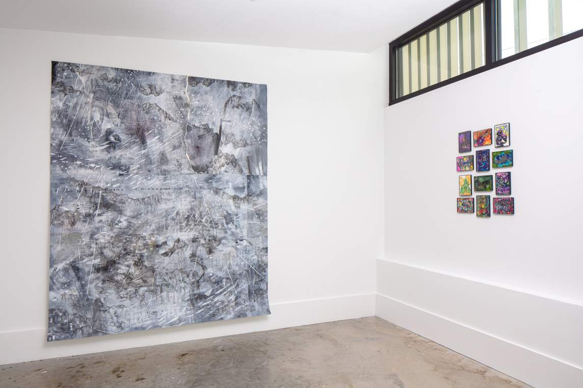 A large black, white, and gray abstract drawing hangs on a white wall, with a grid of smaller abstract works hung on the wall next to it.