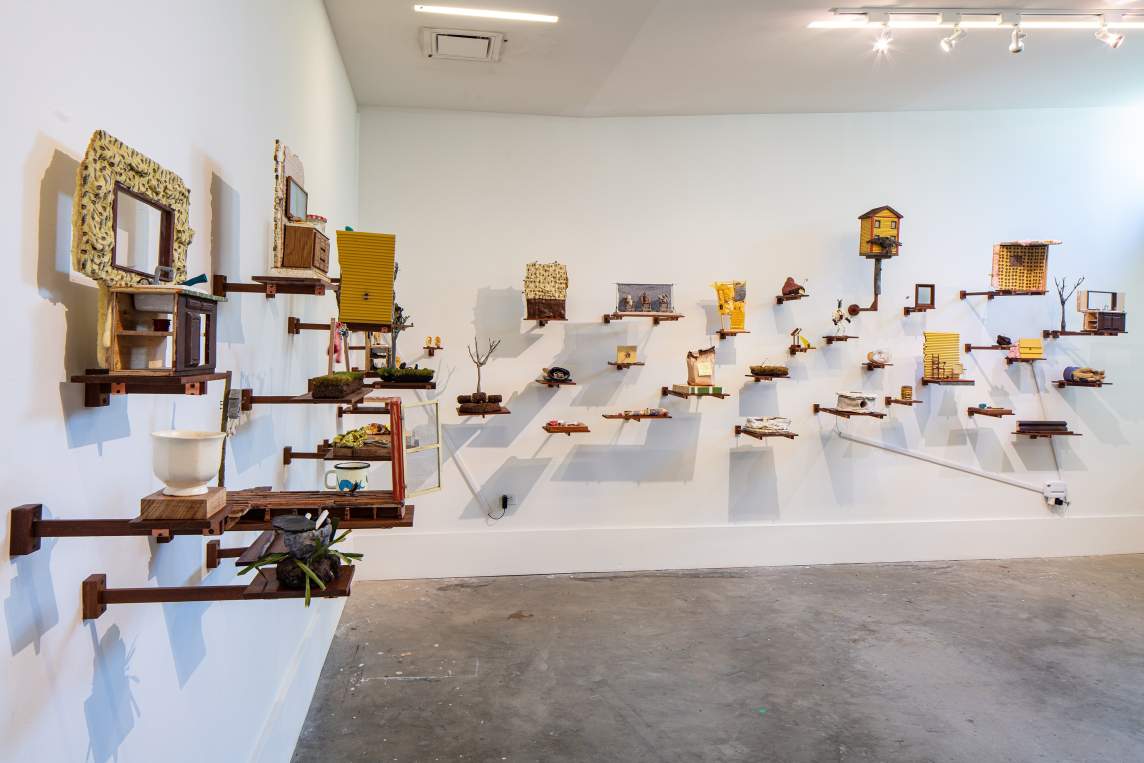 In a room with white walls and concrete floors, dozens of miniature sculptures depicting household objects are hung on brackets to create an art installation.