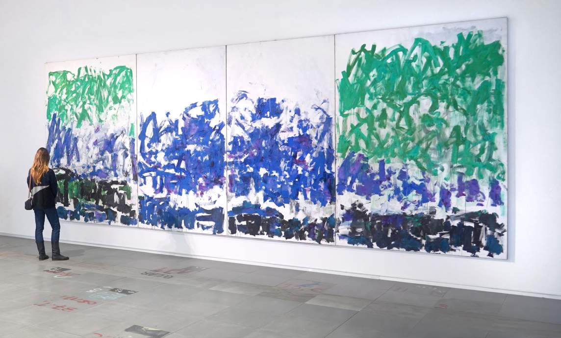 A person observes a large, abstract quadriptych painting by Joan Mitchell, with energetic fields of green, blue, and black brushstrokes on a white background.