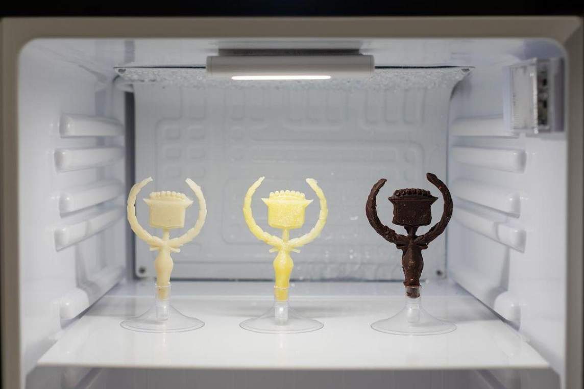 In the interior of a freezer compartment, 3 Cadillac hood ornaments stand in a row on small plastic stands, from left to right, they are made from Shea butter, butter, and chocolate.