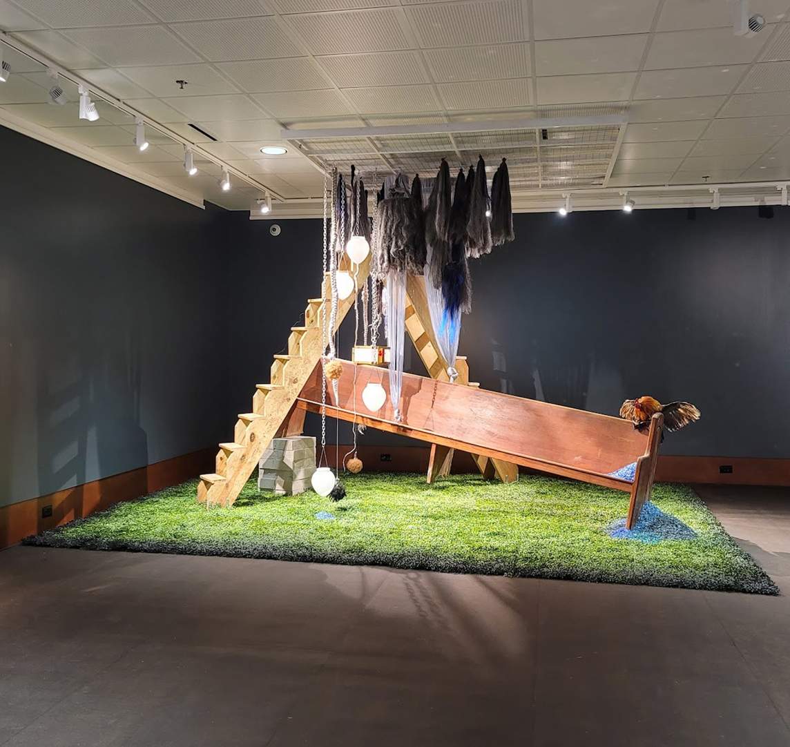 Conterminous Elegies is a mixed media installation. On top of a large square of artificial turf, two wooden stair ladders rise to the ceiling forming a V, and a church pew sits at an angle beneath. Large tassels of synthetic hair and teardrop lamps hang from the ceiling.