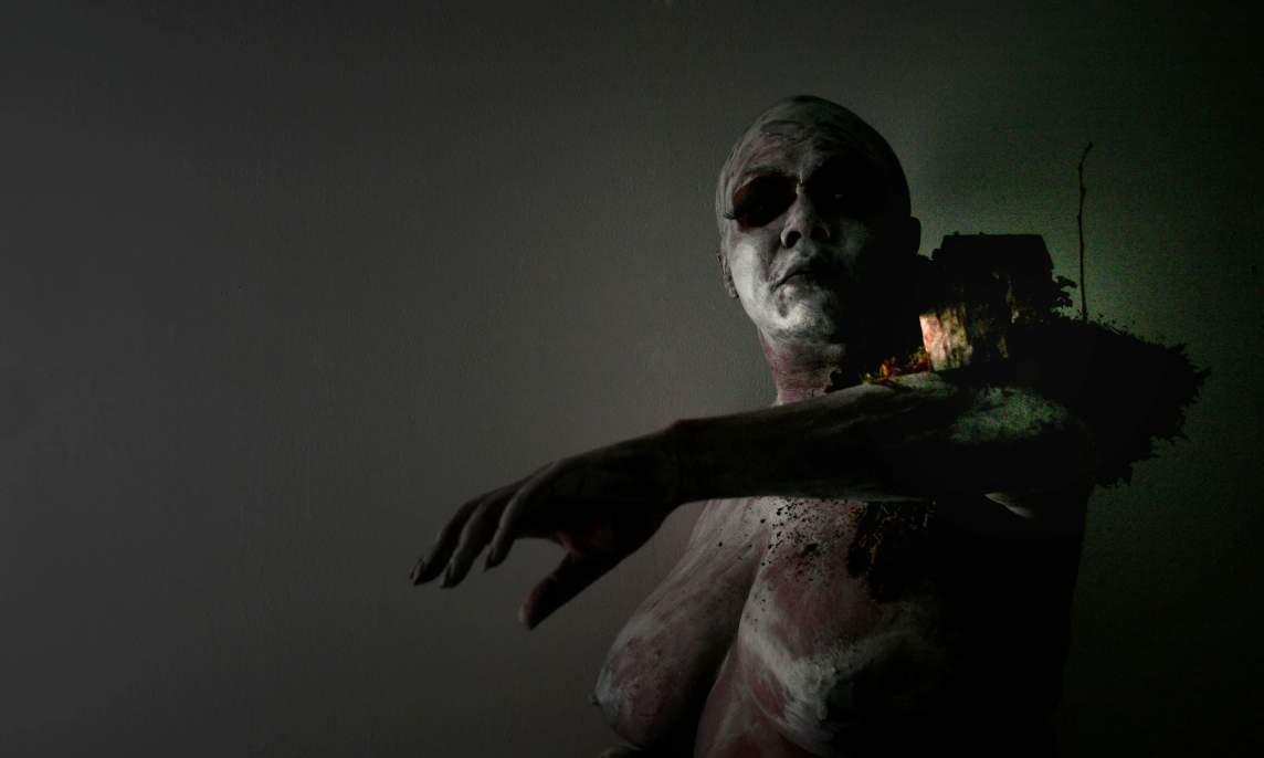 In this darkly lit photo, we see a nude woman from the waist up, with one arm outstretched and a small house-like structure perched on her shoulder in a nest of moss and grass. The woman, who has dark skin partially covered with white paint, looks up over her shoulder at the structure , which is partially lit from within.