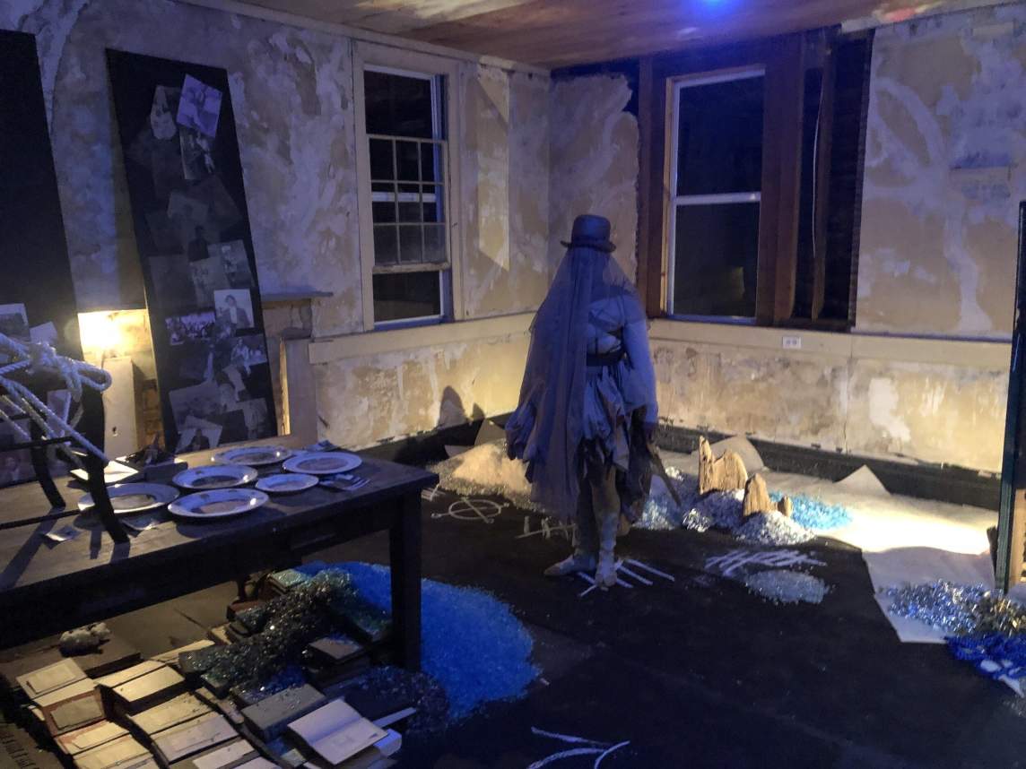In a darkly lit room with distressed plaster walls and windows, a ghostly figure stands, wearing white layers, a veil, and a top hat. Piles of glittery materials can be seen on the floor, along with white chalk drawings of symbols.