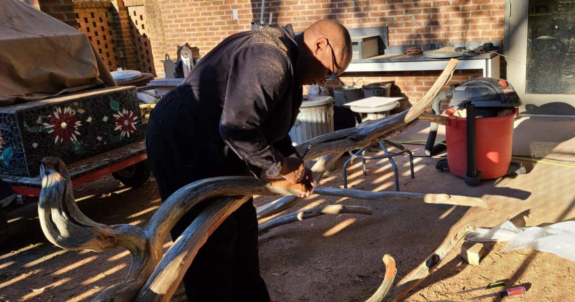 Michaela Pilar Brown in a dark jump suit is working with a large, twisted piece of wood outdoors, with tools and equipment visible in the background.
