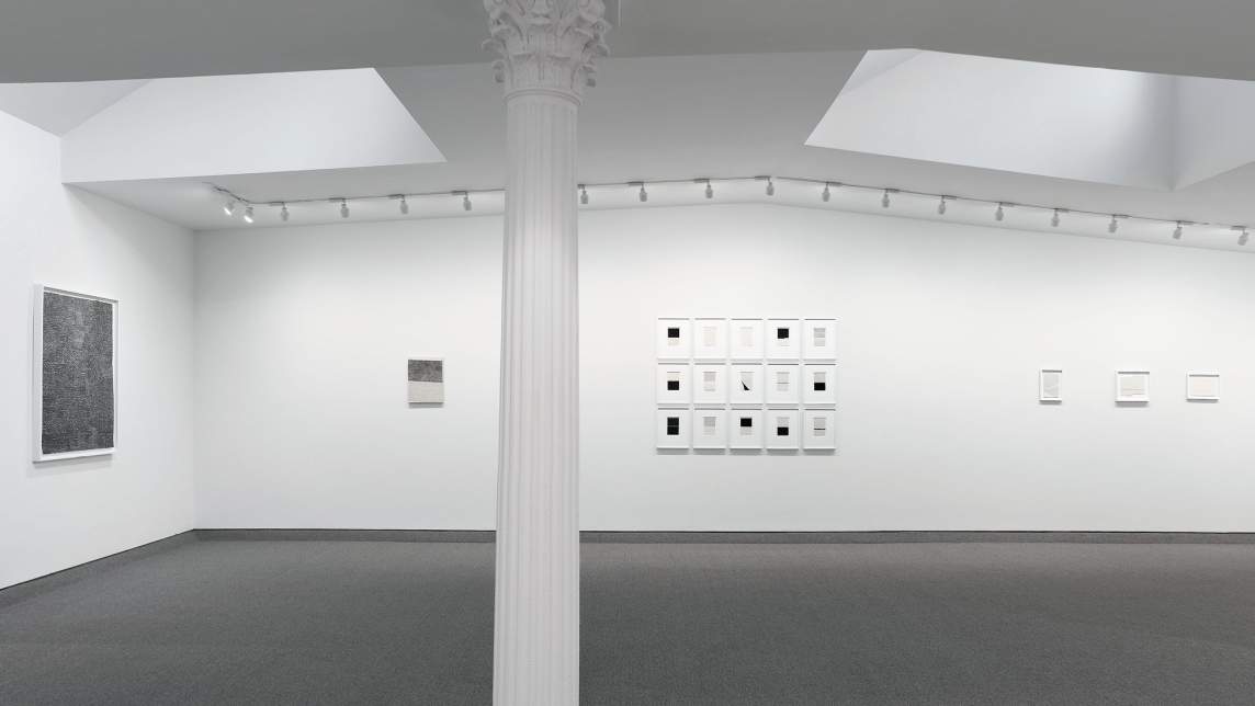 In an art gallery with white walls, sky lights, grey floors, and an ornate central pillar, minimalist black-and-white drawings and prints hang on the walls in white frames.