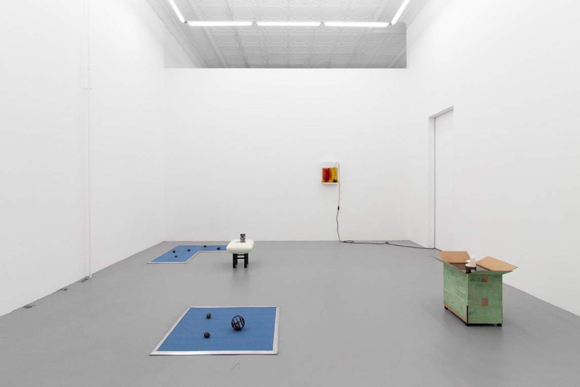 In an art gallery with white walls and gray floors, sculptures by Peggy Chiang are displayed in a spare installation. We see two blue mats on the floor with balls on them, a cardboard box with green painting on the exterior, a wall mounted sculptural box with a red interior, and a free-standing sculpture with a white pillow attached to a stool.