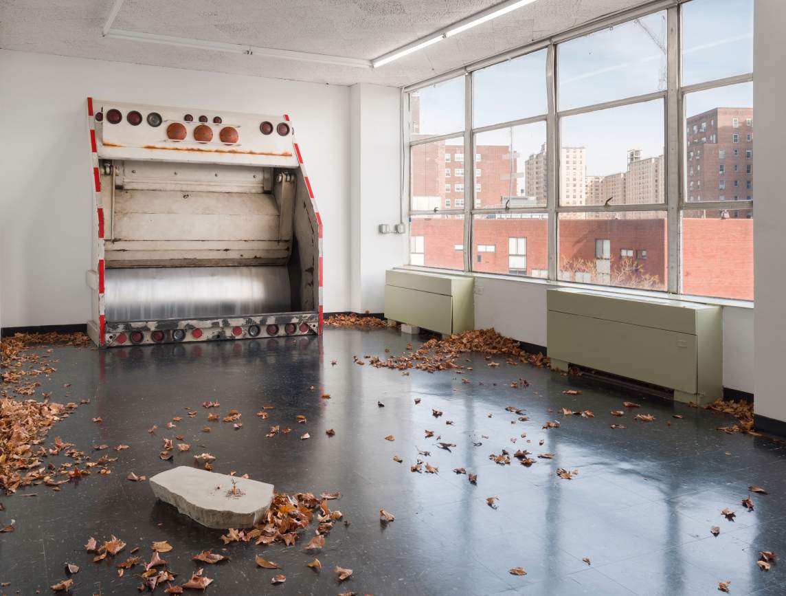 Toss in the asphalt is an installation in a white-walled room with large windows - a reconstruction of the back of a garbage truck stands upright against the wall. Fall leaves are scattered around the floor, piling up at the edges of the walls and in corners as if there’s wind, and a few feet from the truck is a small concrete fragment with a single cigarette burning upright.