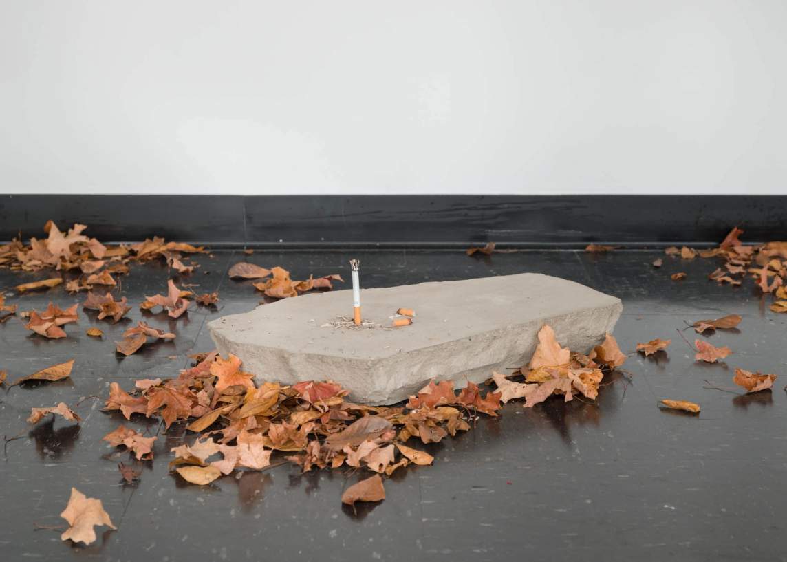 A detail of an art installation shows a chunk of concrete with an upright cigarette sticking out of it. Browned leaves litter the floor around it.