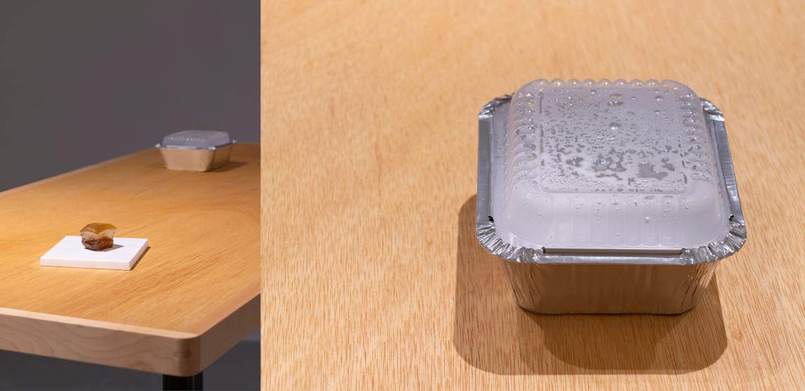 On a table, a takeout container rests with contents obscured by condensation inside. On the other end of the table, a replica of a small hamburger rests on a white square.