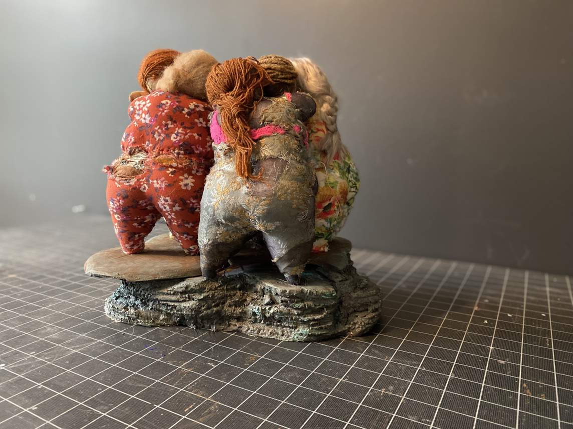 Arc of Affinity is a soft sculpture composed of a group of female figures leaning together in embrace. Their bodies are constructed of remnants of patterned fabric, and they have yarn hair.