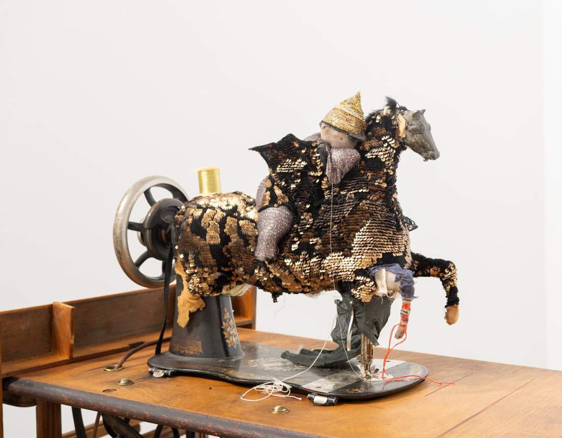 The World is a Loose Stitch is a sculpture that transforms an antique Singer sewing machine resting on a wooden table into figure of a horse and rider. The horse is created with gold and black sequined fabric, and the figure is rendered in silver sequined fabric, with a pointed gold sequined hat. The figure appears to be sewing on the fabric that creates the horse form.