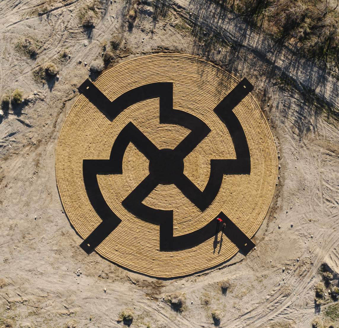 Aerial view of art installation titled Immersion, which is a circular medallion with a black symmetrical, labyrinth-like design on concentric arcs, with a person in red standing near the edge and sparse vegetation surrounding the area.