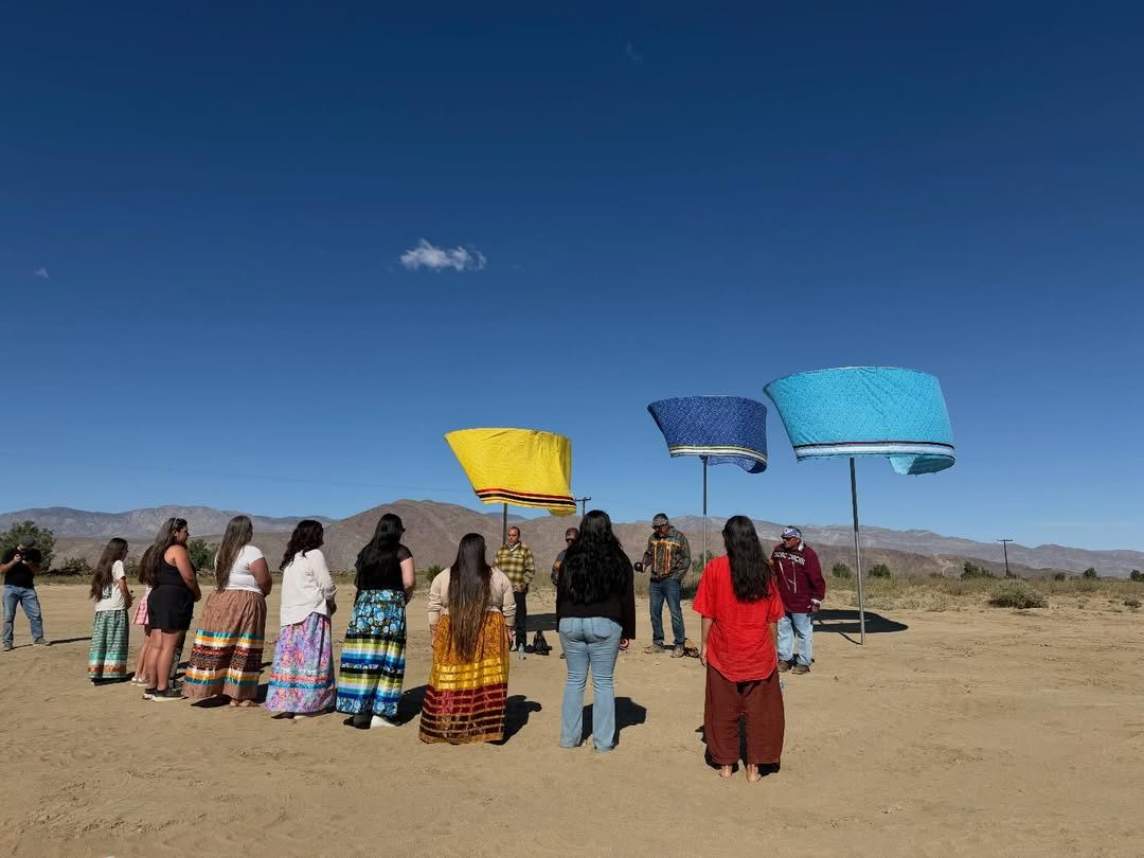 Thirteen men and women stand under or in a row in front of three works of art that consist of fabric banners atop metal poles set up in a sandy desert expanse. Each banner is formed into a ring like a lampshade, and is outlined against a clear, vivid blue sky.
