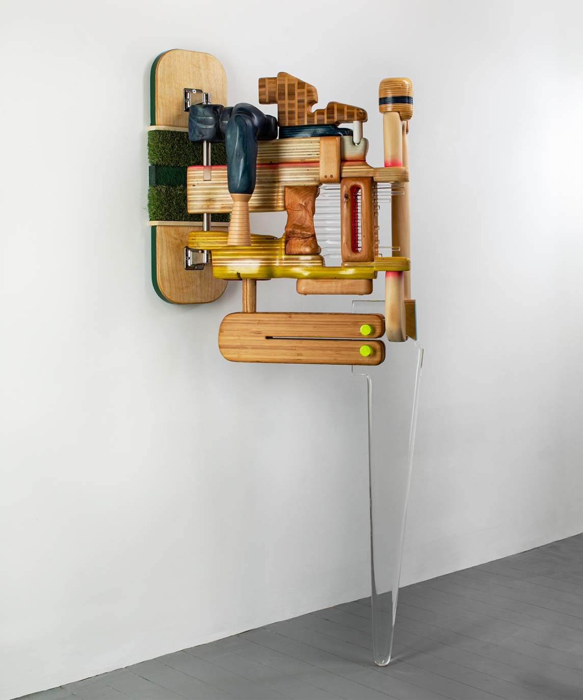 Wood grain is visible on most of the puzzle-like pieces making up a sculpture that hangs on a wall and is supported at its front by a clear Plexiglas leg that tapers to a rounded point where it sits on the gray gallery floor.