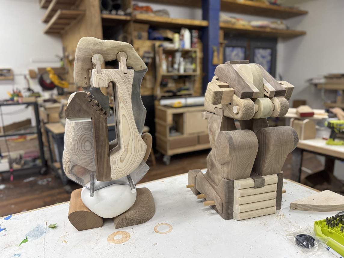 Two wood sculptures sit on a work surface in a studio. Both are made up of light and dark unstained wood carved into rounded or angular, interlocking forms.