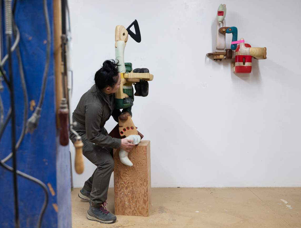 The artist stoops to adjust a sculpture on a wooden base. If she were to stand, the base would be about knee-high and the sculpture would about the same height as the artist. The work is made up of interlocking wooden pieces, some showing the wood grain and others painted in shades of green, yellow, and black.