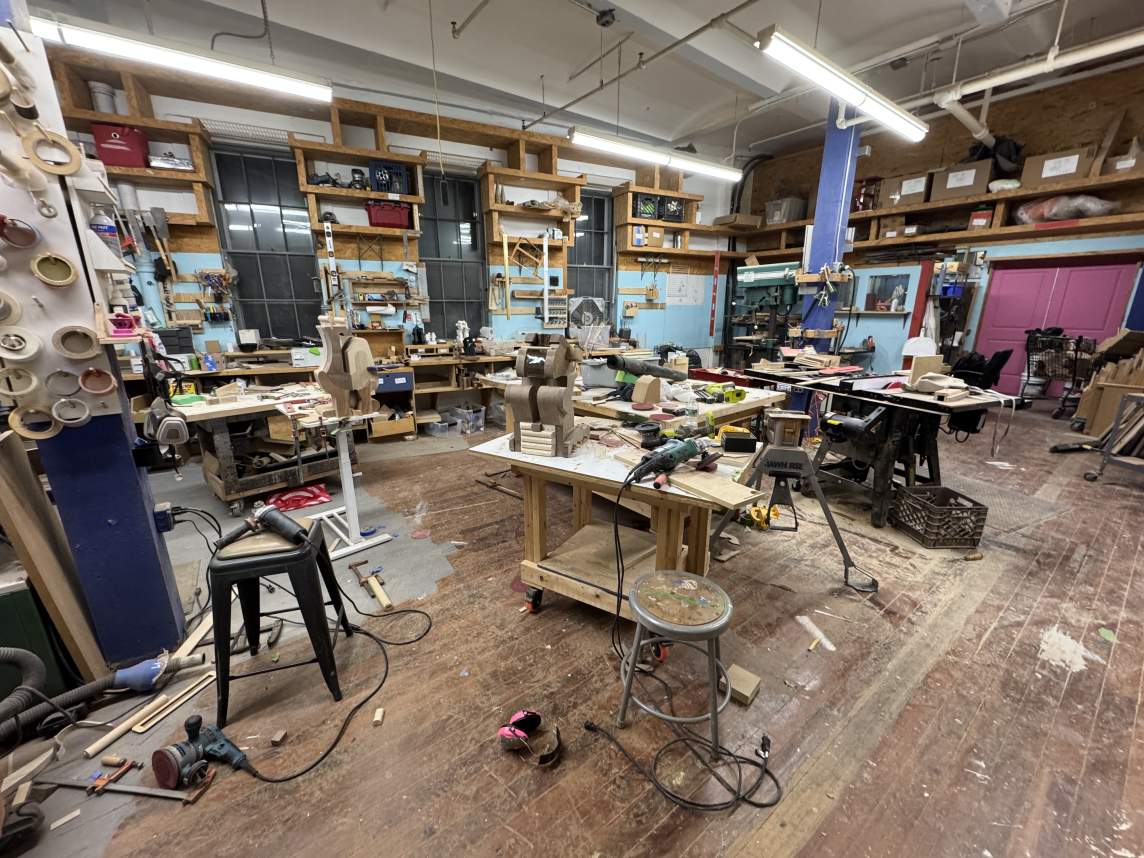 Two wood sculptures made up of rounded, organic shapes are encased in two wood boxes, which perch on two work tables. A drill, nail gun, carving materials, a rubber mallet, a bottle of glue, and other tools lie jumbled around the closer crate, and the second is set against a wall under shelves lined with boxes and near a drill press the height of a person.