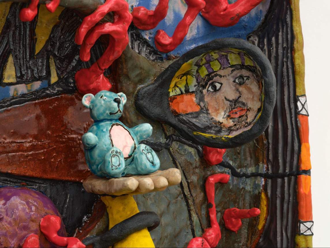 In close detail of ceramic sculpture, a rear view mirror shows a portrait of a man's face with goatee and beaniie, and to left a hand extends holding a turquoise stuffed bear.