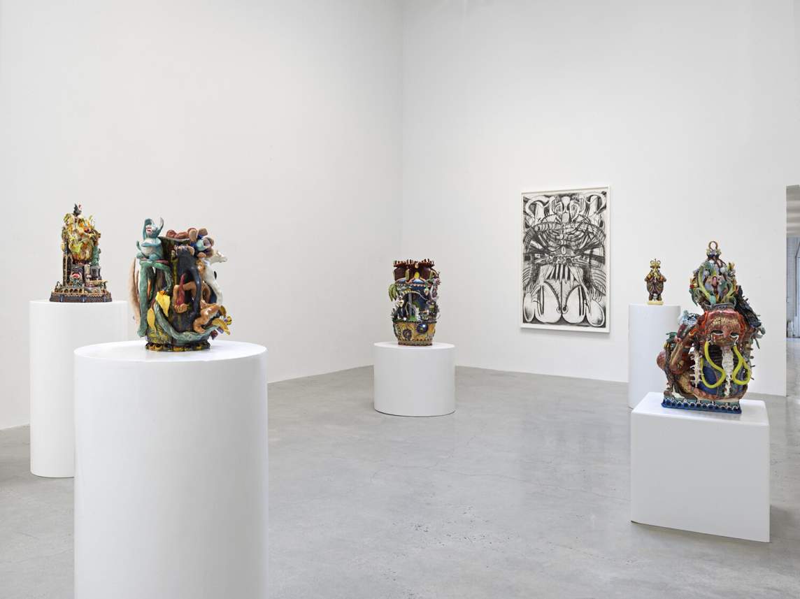 A white gallery room displaying intricate colorful ceramic sculptures on white pedestals and a large black-and-white charcoal drawing on the wall.