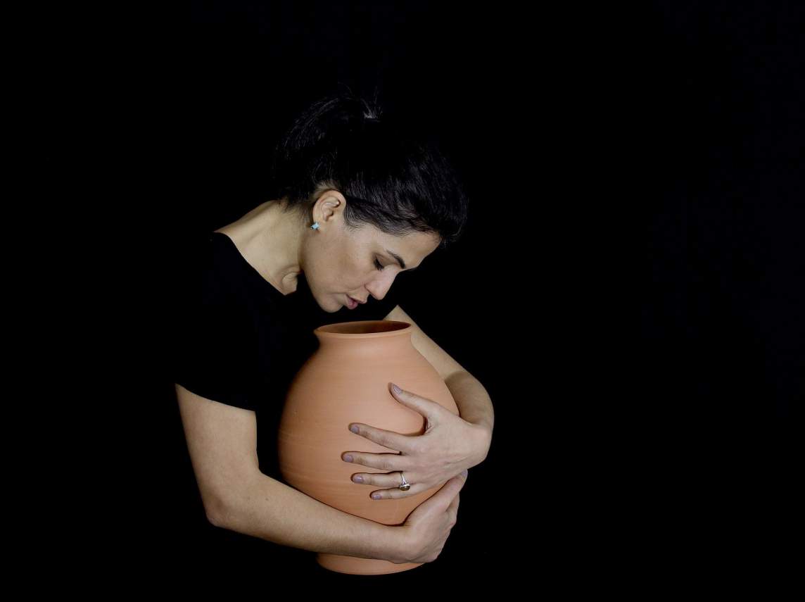 Raheleh Filsoofi, an Iranian-American woman with medium skin tone and dark hair, hugs a terra-cotta orange clay vessel, leaning her head down to whisper names into the open mouth of the pot. She wears a black shirt that disappears in the black background.