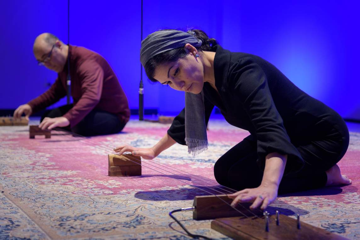 Raheleh and Reza Filsoofi, two Iranian-American people, kneel on a Persian carpet, playing a four-stringed instrument that rests on the floor.