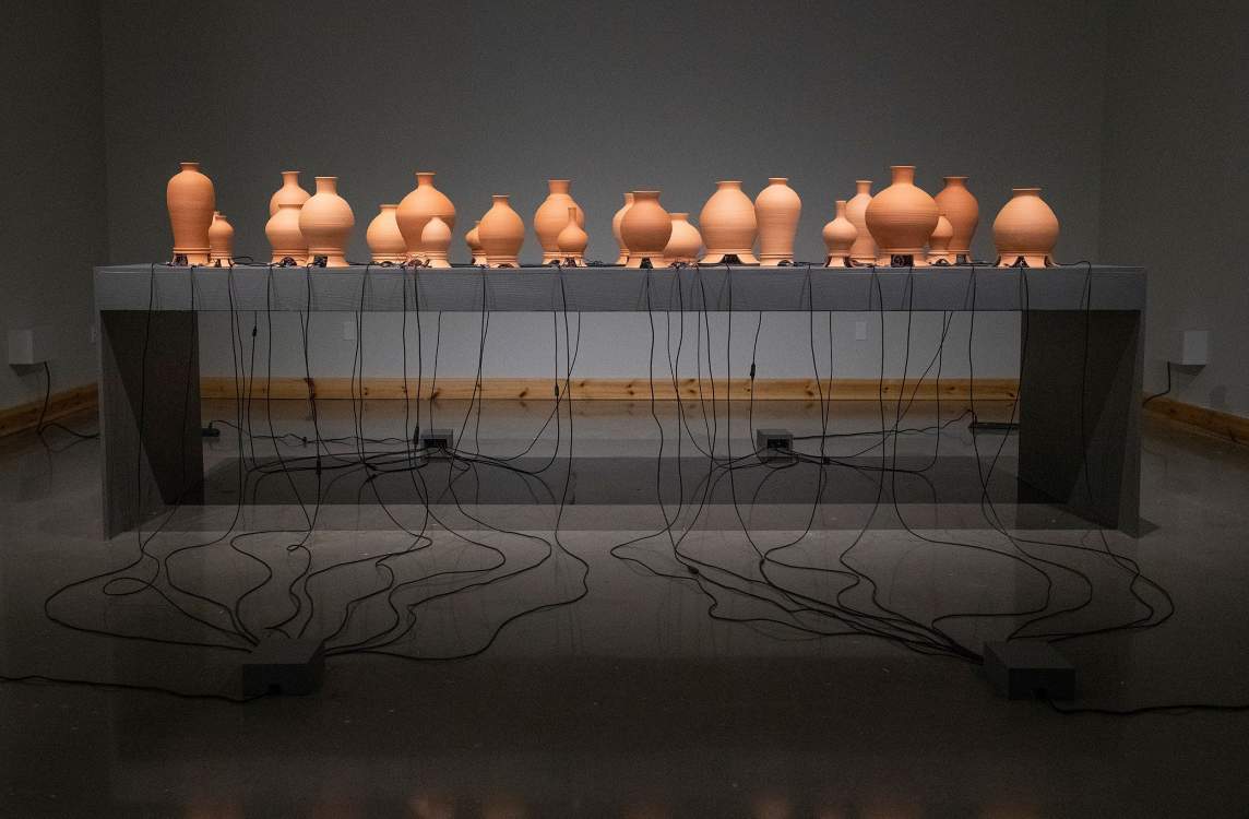 “The Inh(a/i)bited Space” is a sculptural installation of 64 bulbous clay vases of various scales staggered along the length of a long gray table. From openings of the base of each, black wires extend and drape down to the floor, where they connect at 4 electrical junctures.