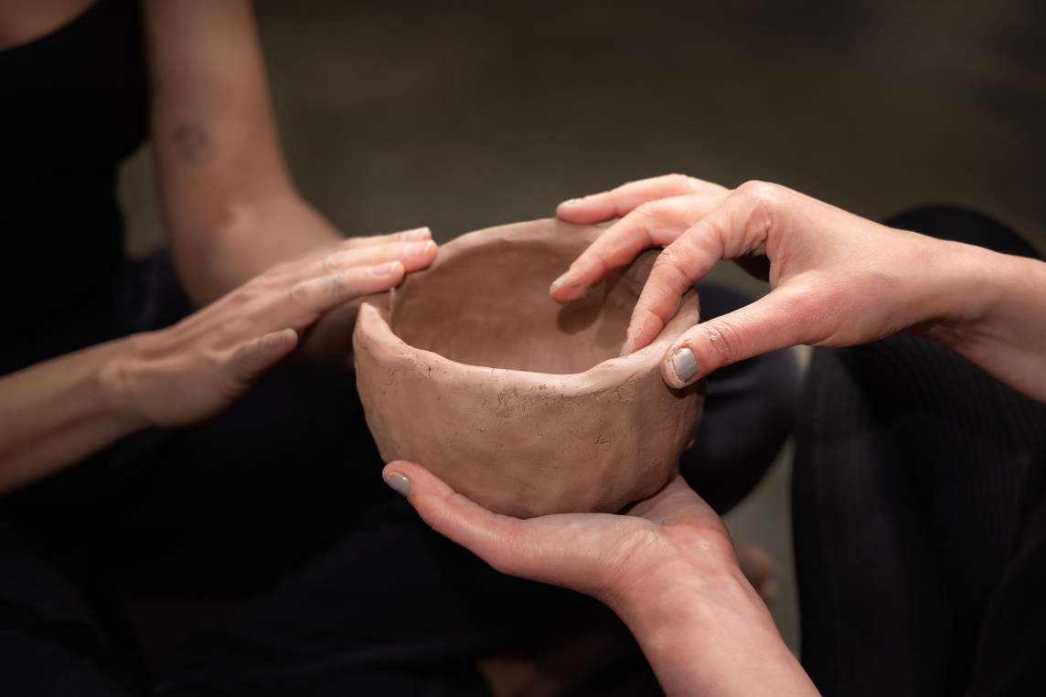 Two sets of hands with medium and light skin tones hold a pinched clay pot that they are forming with their fingers.