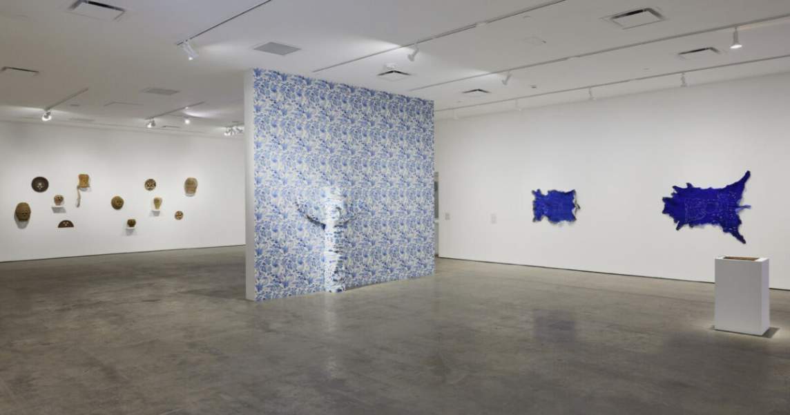 Artworks installed in a gallery space - on the right wall, flattened blue animal hides hang horizontally. In the central area, a short totem pole is wrapped with floral blue toile wallpaper in front of a dividing wall also covered in the wallpaper.