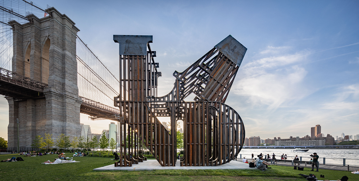 A 30-foot steel sculpture spells LAND in a format reminiscent of Robert Indiana’s 1966 sculpture, LOVE, on the lawn of a park by the water, with a bridge and cityscape in the background. Many people mill about on the lawn.
