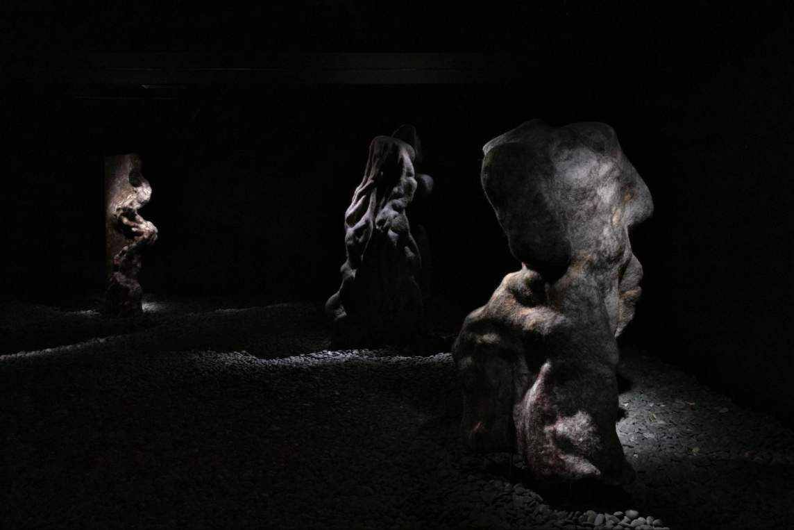 “Olistrostrome” is a sculptural installation in a very dark room, with light cast onto 3 large indistinct woolen biomorphic forms, staggered across the room on a floor of gray river stones. The forms have undulating wrinkles and bulbous extrusions, and smooth faces elsewhere, evoking both figures and rock formations.