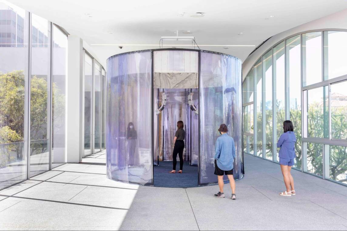 “the input of this machine is the power an output contains” is a multimedia installation, in which a performer is reciting a text generated by artificial intelligence. In this view, a masked person stands inside curved plexiglas walls, and two other people look on from outside the installation.