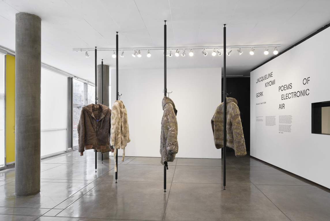 An art installation featuring various textured coats hanging from ceiling-mounted rods in a gallery with white walls, a concrete pillar, and text panels.
