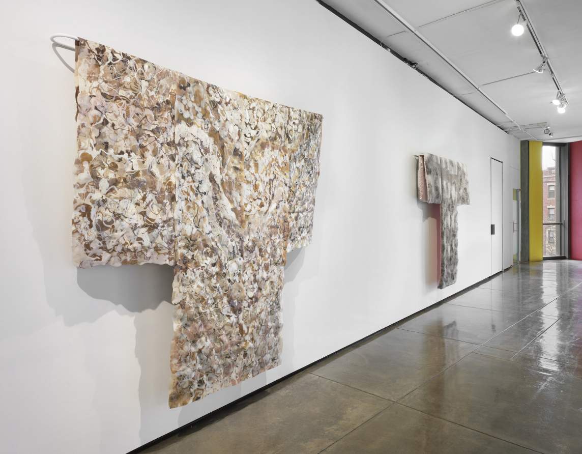 Art gallery interior featuring two fabric sculptures with textured, neutral-colored surfaces, displayed in the manner of Japanese kimono on white walls with a reflective grey floor.