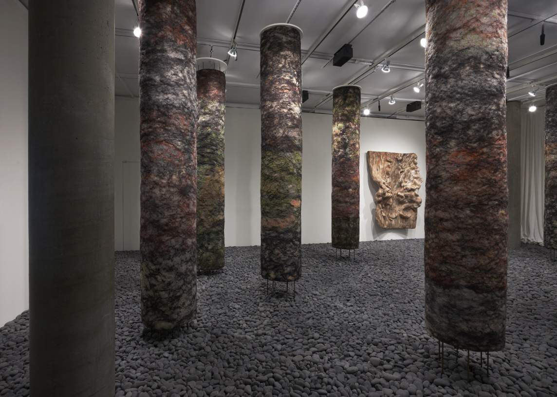 An art installation featuring cylindrical columns with textured, colorful surfaces standing on a floor covered in gray pebbles, located in a gallery with soft lighting.