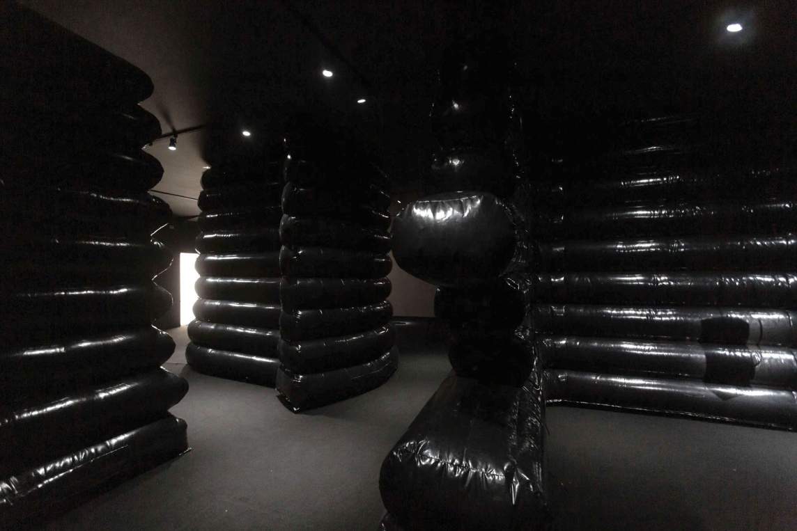 A dimly lit room filled with large, glossy black inflatable structures stacked in layers, creating a labyrinthine atmosphere.