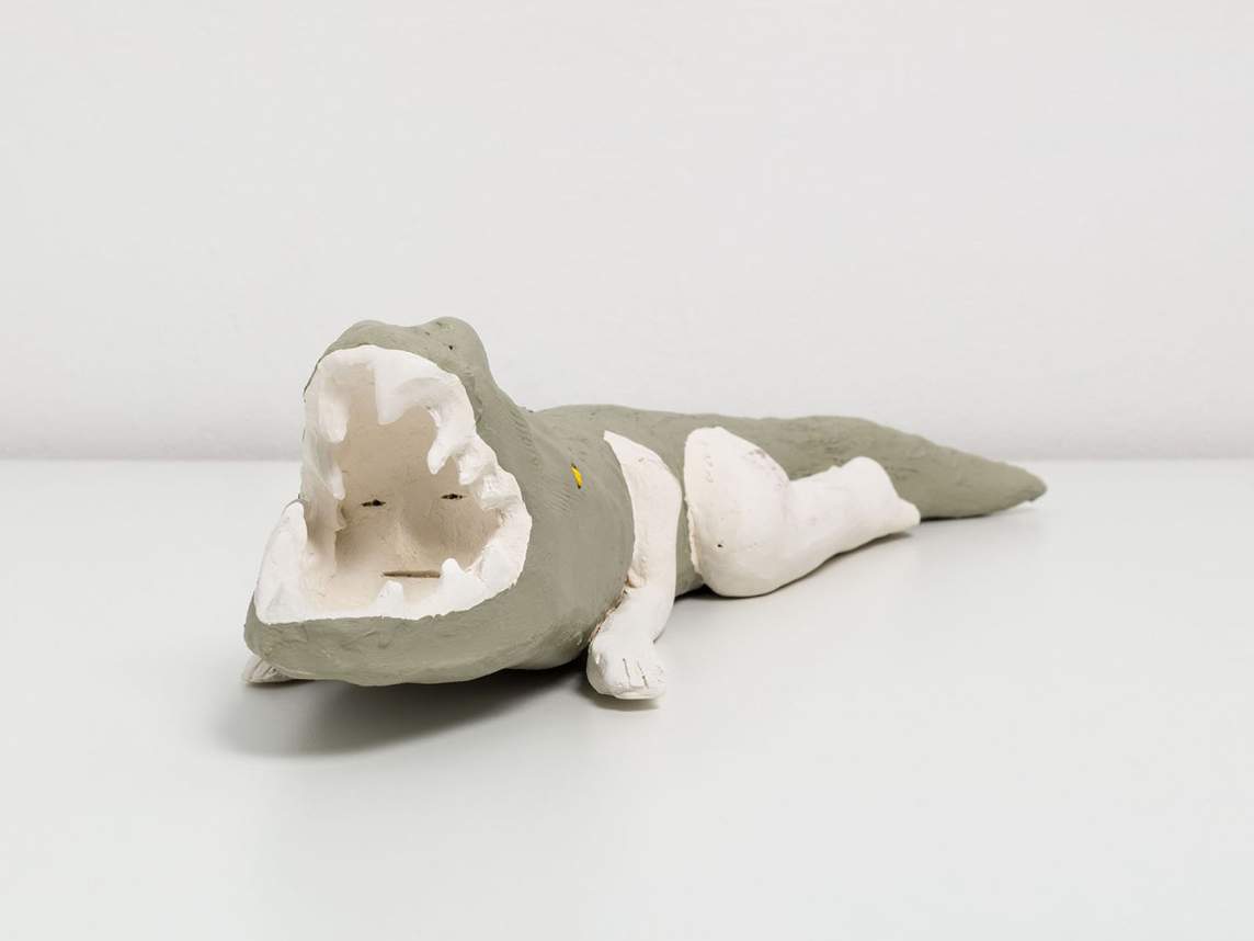 An off white doll sized ceramic sculpture of a woman with a dog’s head, and dog nipples lying down on her back. Her eyes are carved out from the clay to look like closed eyes with long eyelashes. Her left hand is resting on her left thigh, and her right hand rests flat against her right leg.