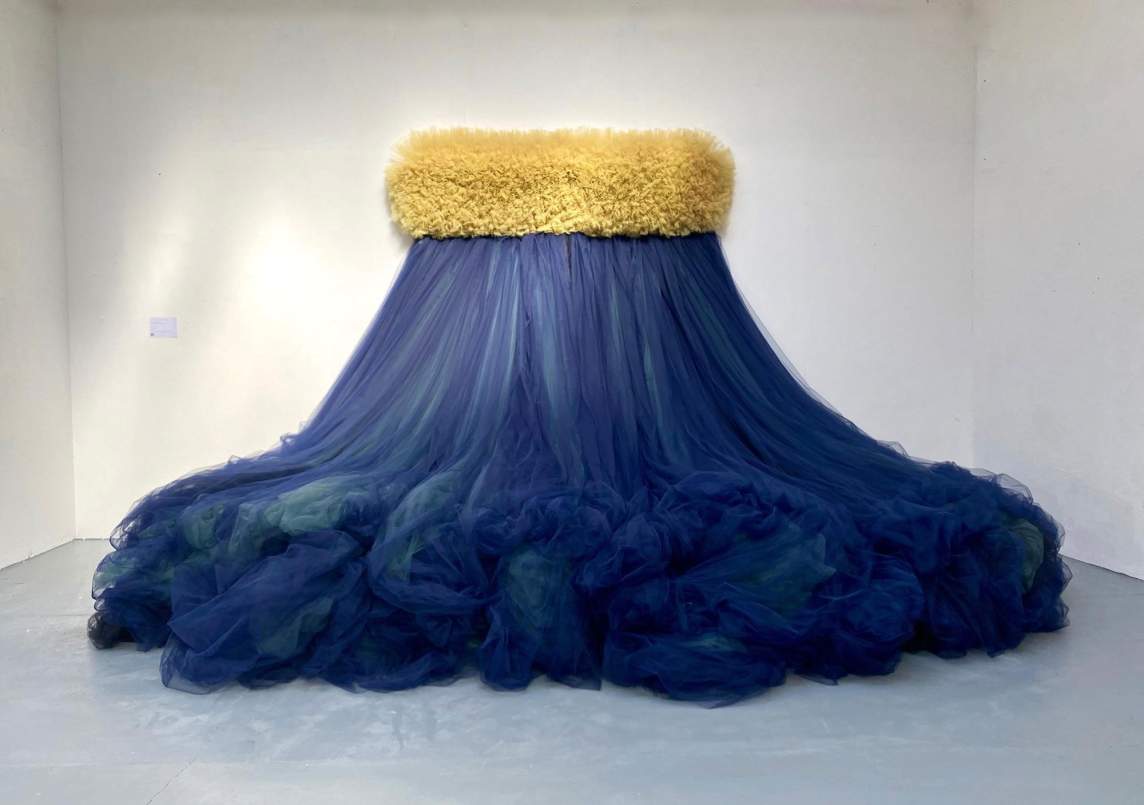 “Only the Air Was Heard” is a sculptural installation on the white wall of a museum space. From a rounded rectangular band of densely ruffled mustard tulle on the wall, a cascading form of bundles of gray and lavender tulle fabric spill down, piling outwards onto the floor below.