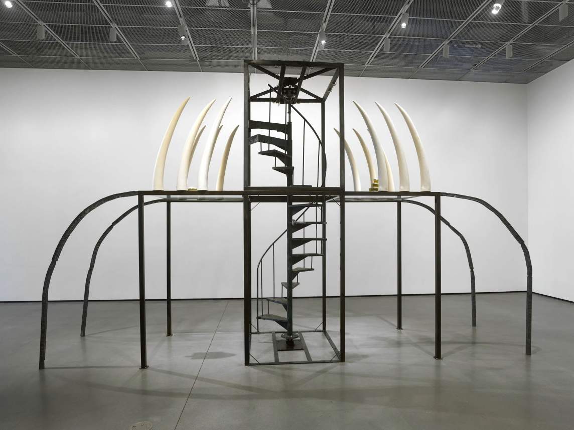 This spidery sculpture stretches four thin black legs out long, curving down to meet the gray floor. Another set of four legs, these straight, support a central frame, which encloses a black metal spiral staircase that corkscrews up the center. White tusk-like forms sit in rows on the second level, all curving up and inward.
