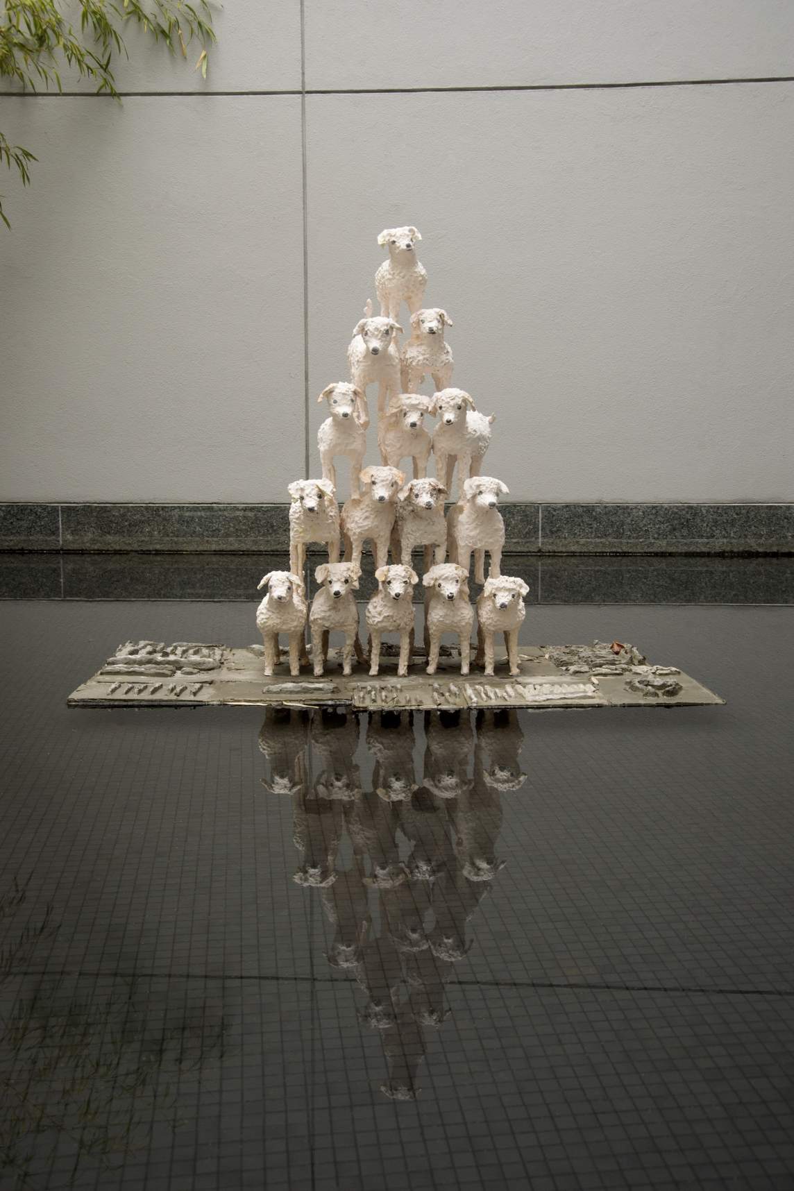 Untitled (15 Esthers in a pyramid) is a sculpture of 15 white poodles made of mottled clay stacked in a cheerleader-like pyramid formation. They stand on a rectangular cement plinth with arranged objects above a dark reflective pool.